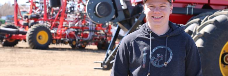 Student wearing a hoodie and hat, standing in a lot with red farming equipment